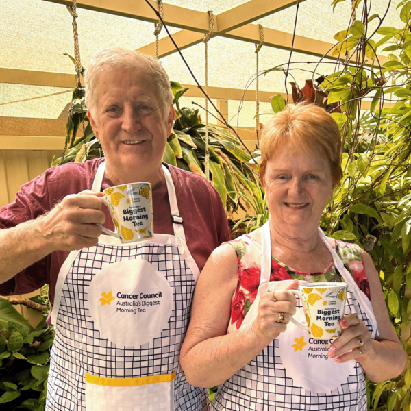 A man and a woman pose to camera holding Biggest morning tea mugs and wearing Biggest morning tea cooking aprons